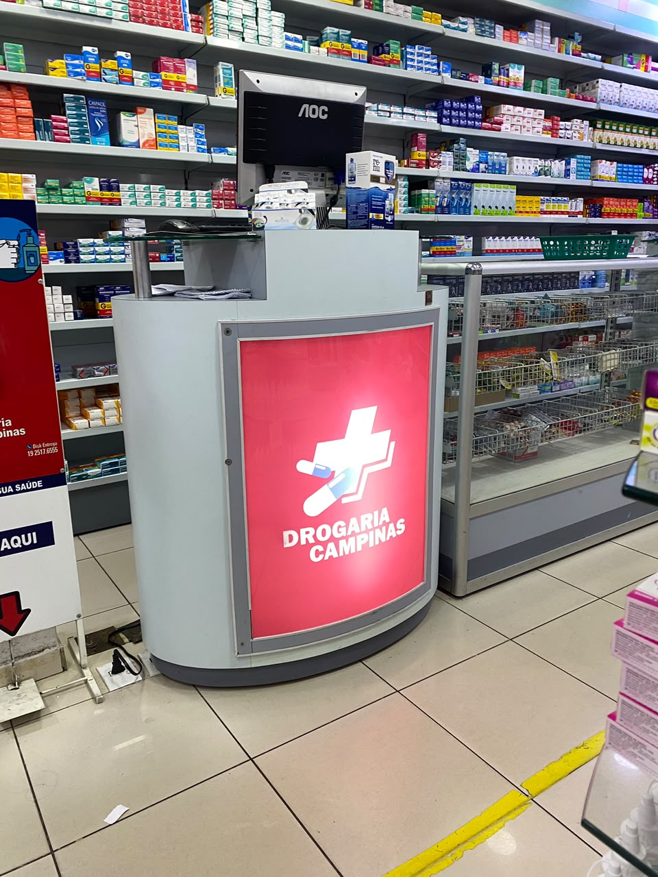 Balcão de atendimento da Drogaria Campinas com a marca da farmácia e prateleiras de medicamentos